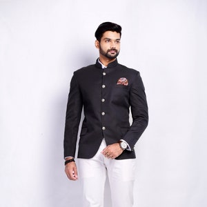 Puede incluir: Una chaqueta negra con cuello mandarín y cierre de botones. La chaqueta tiene un bolsillo en el lado izquierdo con un pañuelo de bolsillo estampado. El modelo lleva pantalones blancos y zapatos de cuero marrón.