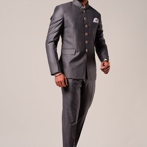 K&ouml;nnte beinhalten: Ein anthrazitfarbener Anzug mit einer Jacke mit Stehkragen und passender Hose. Die Jacke hat goldfarbene Kn&ouml;pfe und ein Einstecktuch. Das Outfit wird mit braunen Lederloafers kombiniert.