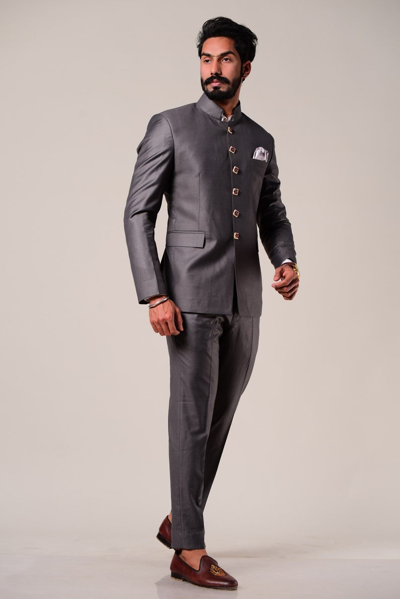 K&ouml;nnte beinhalten: Ein anthrazitfarbener Anzug mit einer Jacke mit Stehkragen und passender Hose. Die Jacke hat goldfarbene Kn&ouml;pfe und ein Einstecktuch. Das Outfit wird mit braunen Lederloafers kombiniert.