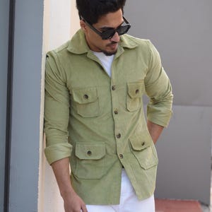 Könnte beinhalten: Ein Mann trägt ein hellgrünes Cordhemd mit mehreren Taschen und ein weißes T-Shirt darunter. Er trägt eine weiße Hose und eine schwarze Sonnenbrille.