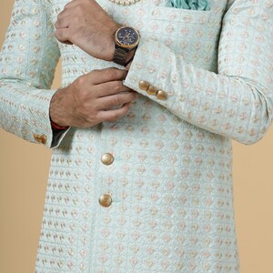 May include: A light blue embroidered jacket with gold buttons. The jacket has a quilted pattern and a pocket on the left side. The person is wearing a pearl necklace and a watch.