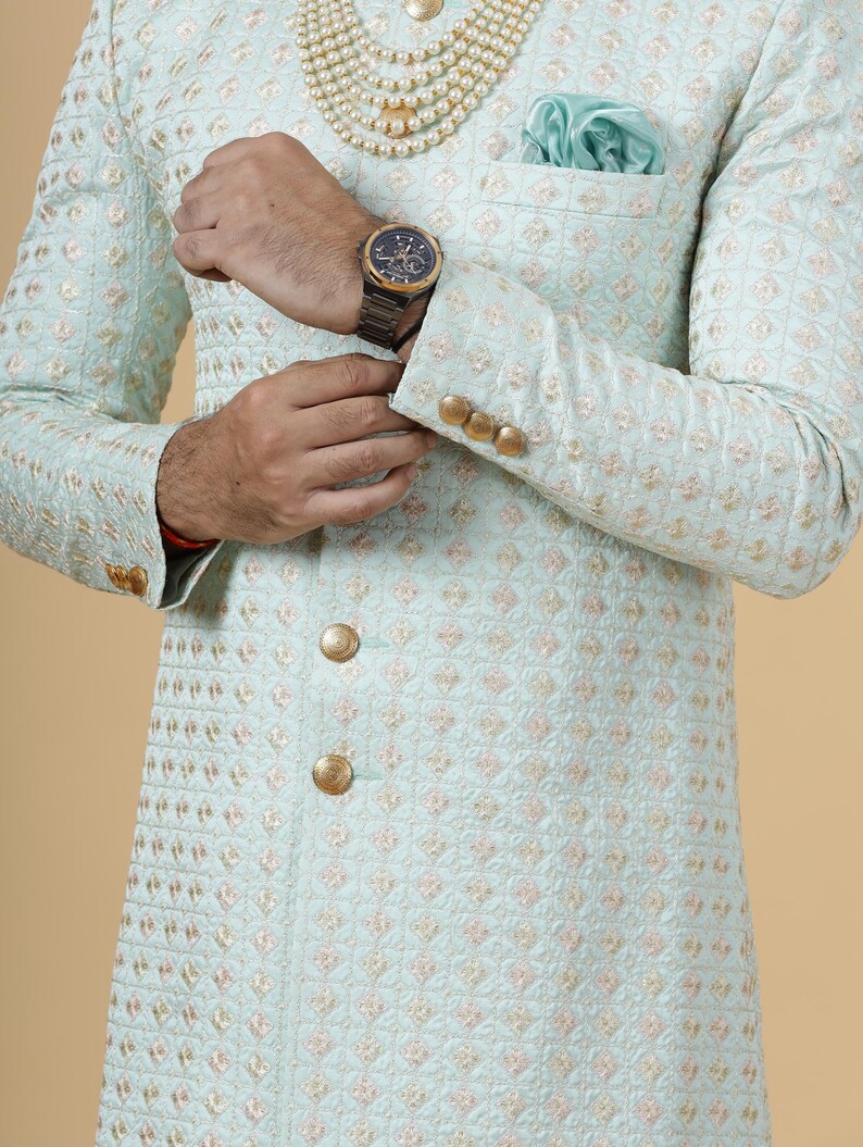 May include: A light blue embroidered jacket with gold buttons. The jacket has a quilted pattern and a pocket on the left side. The person is wearing a pearl necklace and a watch.