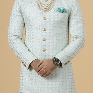 May include: A light blue patterned men's sherwani with gold buttons and a pearl necklace. The sherwani has a pocket on the left side with a light blue handkerchief.