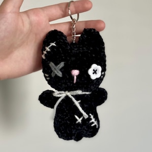 May include: A black crochet cat keyring with white stitching, a grey cross, a pink nose, and a white button eye. The cat has a white bow and is attached to a silver keyring.