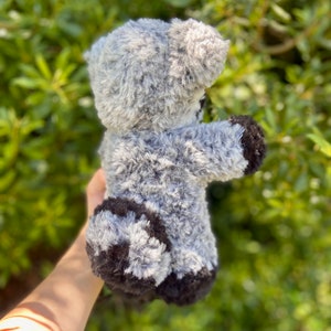 May include: A gray and black plush teddy bear with a soft, fuzzy texture. The bear has a light gray body with darker gray and black accents on its paws and ears. The toy is held up against a backdrop of green foliage.