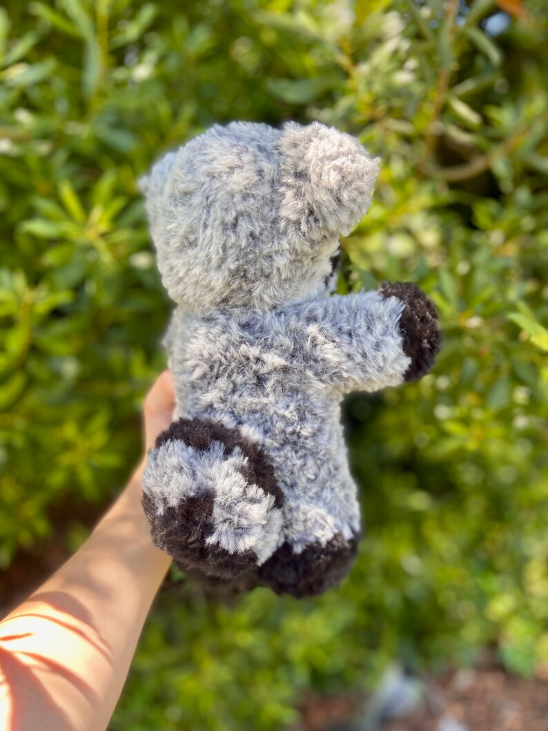 May include: A gray and black plush teddy bear with a soft, fuzzy texture. The bear has a light gray body with darker gray and black accents on its paws and ears. The toy is held up against a backdrop of green foliage.