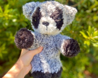 Könnte beinhalten: Ein Plüsch-Waschbär-Stofftier mit grau-weißem Körper, schwarzen Akzenten an Pfoten, Ohren und um die Augen. Das Kuscheltier wird vor einem verschwommenen grünen Hintergrund gehalten.