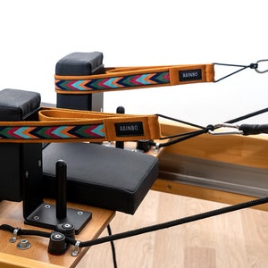 May include: Two black padded blocks with straps attached to a wooden pilates reformer. The straps are orange, blue, green, and red with the word "RAINBO" printed on them.