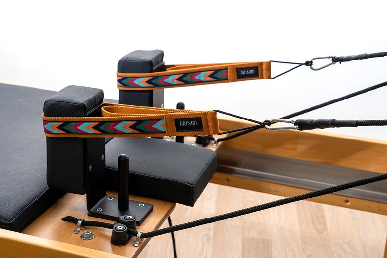 May include: Two black padded blocks with straps attached to a wooden pilates reformer. The straps are orange, blue, green, and red with the word "RAINBO" printed on them.