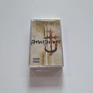 May include: A cassette tape with a white and brown background. The text "Devil Driver" is printed in black on the front of the tape. A parental advisory sticker is in the bottom left corner.