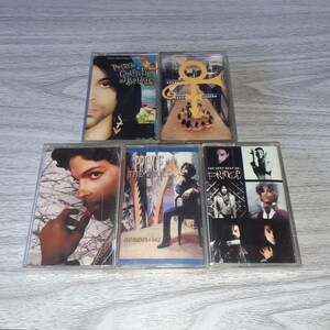 May include: A collection of six cassette tapes featuring Prince. The tapes are in clear plastic cases with colorful artwork. The titles of the tapes are "Prince Graffiti Bridge", "The Vault", "Old Friends 4 Sale", "The Very Best of Prince", and "Purple Rain".