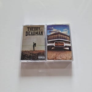 Peut inclure: Deux cassettes audio du groupe Theory of a Deadman. La cassette de gauche présente une image en noir et blanc d'un homme avec des ailes debout dans un champ. La cassette de droite présente une image couleur d'une voiture ancienne avec une plaque d'immatriculation qui indique "Theory of a Deadman".