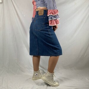 May include: A dark blue denim skirt with a high waist and a button closure. The skirt is knee-length and has a straight silhouette. The skirt is worn with a white and red gingham top and white high-top sneakers.