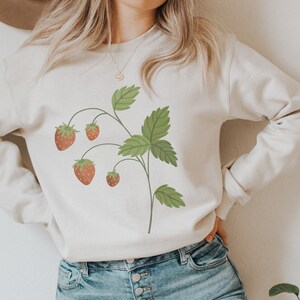 Könnte beinhalten: Ein weißes Sweatshirt mit einem grünen Erdbeerpflanzen-Motiv. Die Pflanze hat fünf rote Erdbeeren und grüne Blätter.