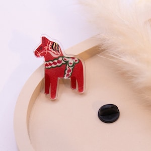 Könnte beinhalten: Ein roter Dala-Pferde-Ansteckpin mit weißen und grünen dekorativen Akzenten. Der Pin besteht aus klarem Acryl und hat einen schwarzen Pin-Rücken. Das Pferd ist ein traditionelles schwedisches Volkskunstsymbol.