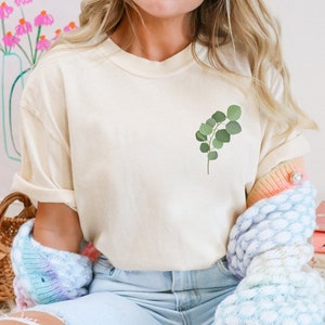 Könnte beinhalten: Ein cremefarbenes T-Shirt mit einem kleinen grünen Eukalyptus-Zweig-Motiv auf der linken Brust.
