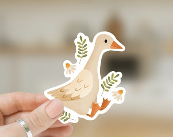 Könnte beinhalten: Ein Aufkleber mit einer Aquarellillustration einer Gans mit beigem Körper, orangefarbenem Schnabel und Füßen sowie weißen Details. Die Gans ist von grünen Blättern und weißen Blumen umgeben. Der Aufkleber wird von einer Hand gehalten.