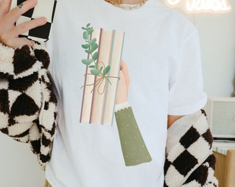 May include: White t-shirt with a graphic of a hand holding a stack of books tied with twine and a sprig of green leaves.