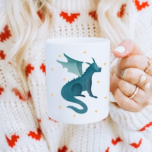Könnte beinhalten: Eine weiße Keramiktasse mit einer blauen Drachenillustration und goldenen Sternen. Der Drache hat grüne Flügel und sitzt auf seinem Schwanz.