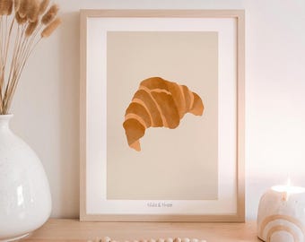 May include: A framed print featuring a watercolor illustration of a croissant in shades of brown and tan. The print is displayed on a light-colored surface, with a vase of dried flowers, a candle, and wooden beads nearby. The text "lido & thee" is at the bottom.