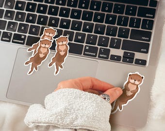 May include: Four brown otter stickers on a silver laptop keyboard. The stickers are in various poses, with one otter sticker being held by a hand.