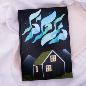 Pode incluir: Um livro preto de capa dura com uma ilustração verde e azul de uma casa com telhado verde e um céu estrelado. A palavra hebraica "Shalom" está escrita no céu em uma fonte estilizada.