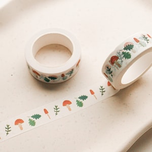 May include: A roll of white washi tape with a repeating pattern of red mushrooms, green leaves, and white stems. The tape is partially unrolled on a white surface.