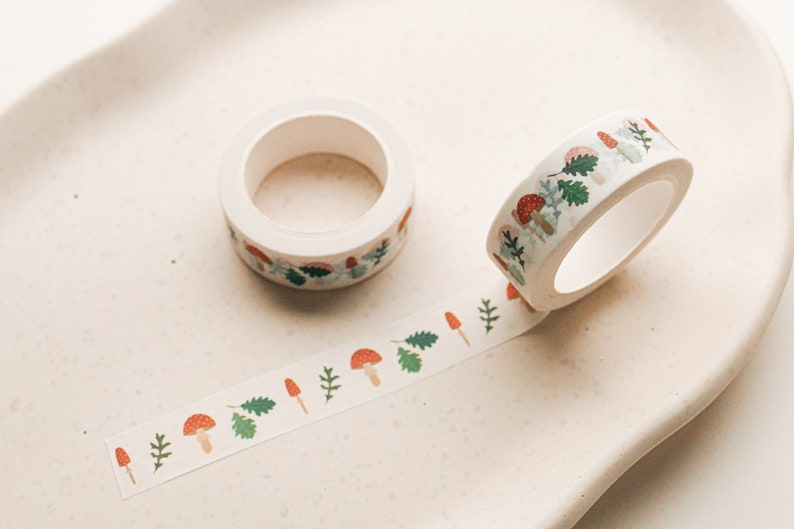 May include: A roll of white washi tape with a repeating pattern of red mushrooms, green leaves, and white stems. The tape is partially unrolled on a white surface.