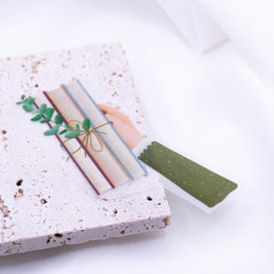 Könnte beinhalten: Ein Stapel Bücher mit einem grünen, belaubten Zweig, der mit Schnur zusammengebunden ist, und einem grünen, gemusterten Washi-Tape-Aufkleber.