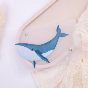 Peut inclure: Une baleine bleue avec un ventre blanc et une petite bouche rose est peinte sur une feuille de papier translucide. La baleine est entourée de petites bulles blanches et de deux petits poissons verts.