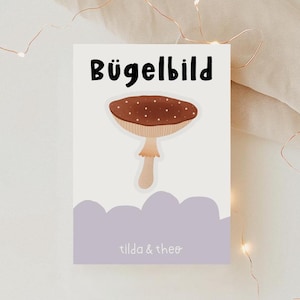 May include: A decorative card featuring a brown mushroom with white spots, set against a light background. The word "Bügelbild" is at the top, with "tilda & theo" at the bottom. The design includes a purple cloud-like shape.