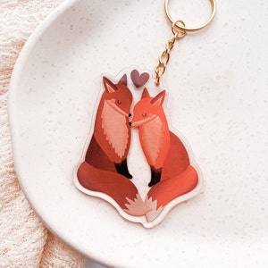 Könnte beinhalten: Ein klarer Acryl-Schlüsselanhänger mit zwei Cartoon-Füchsen in Orange- und Brauntönen und einem kleinen lila Herz darüber. Der Schlüsselanhänger hat eine goldfarbene Kette und einen Ring.