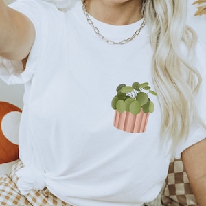 Könnte beinhalten: Weißes T-Shirt mit einem Aufdruck einer grünen Pflanze in einem rosafarbenen Topf.