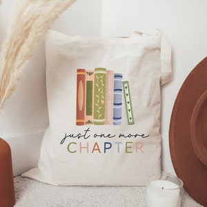 May include: A white canvas tote bag with a colorful illustration of stacked books and the text "just one more CHAPTER".