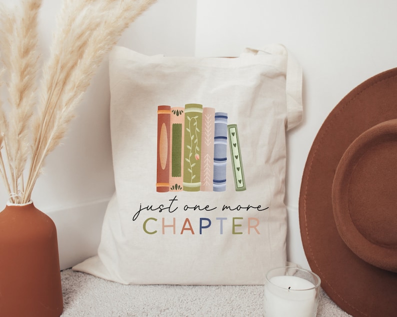 May include: A white canvas tote bag with a colorful illustration of stacked books and the text "just one more CHAPTER".
