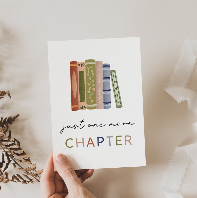 Peut inclure: Une carte blanche avec une pile de livres illustr&eacute;s color&eacute;s et le texte "just one more CHAPTER" en &eacute;criture cursive noire.