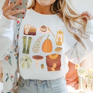Könnte beinhalten: Weißer Pullover mit einem bunten Herbst-Motiv. Das Design beinhaltet Kürbisse, Blätter, Stiefel, eine Laterne, ein Zimtschnecken, einen Pullover und einen Pilz.