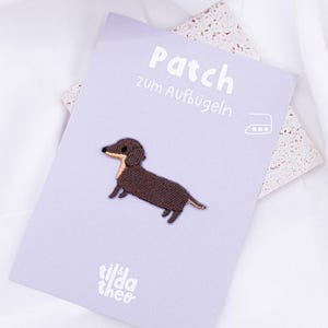 Può includere: Un toppa da stirare a forma di bassotto marrone con il testo "Patch zum Aufbügeln" e il nome del marchio "tilda theo" su uno sfondo viola chiaro.