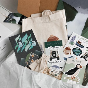 May include: A white gift box filled with various items, including a tote bag, cards, stickers, and a magnet. The box has a label that says "Auf ins n&auml;chste Abenteuer" and "Alles Liebe Hannah". The items in the box have a nature theme with illustrations of houses, birds, and landscapes.