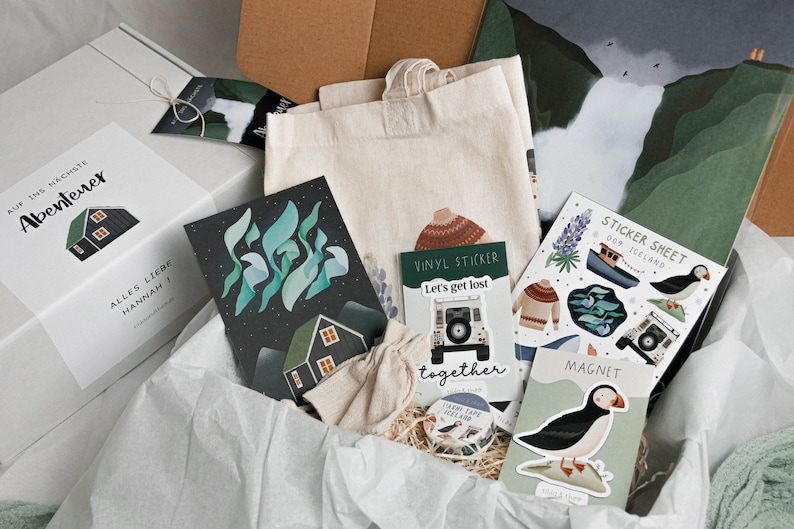 May include: A white gift box filled with various items, including a tote bag, cards, stickers, and a magnet. The box has a label that says "Auf ins n&auml;chste Abenteuer" and "Alles Liebe Hannah". The items in the box have a nature theme with illustrations of houses, birds, and landscapes.