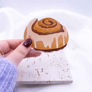 Puede incluir: Un primer plano de una pegatina de rollo de canela. La pegatina es marrón y blanca con un remolino de canela en la parte superior. La pegatina está sobre un fondo blanco.