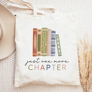 以下が含まれることがあります： カラフルな本の積み重ねと「just one more CHAPTER」というテキストがデザインされた白いキャンバス地のトートバッグです。