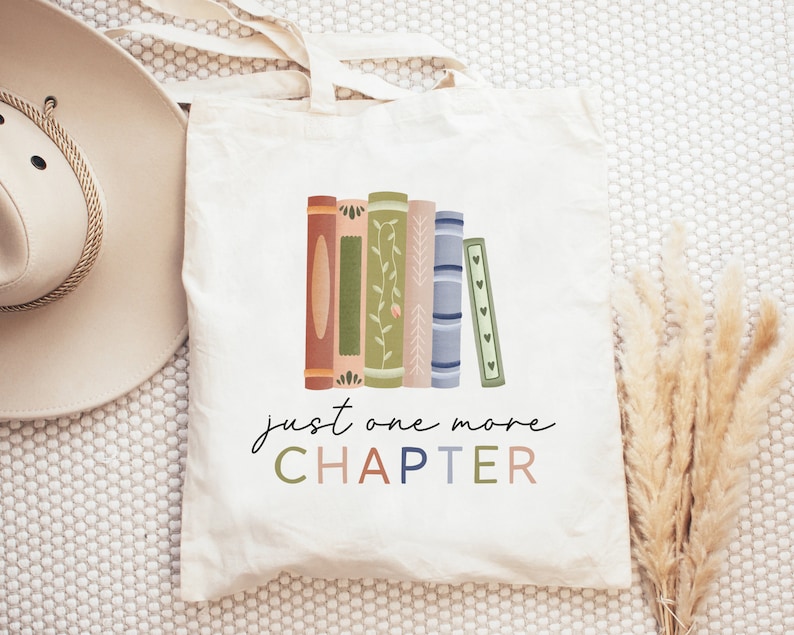 May include: A white canvas tote bag with a design of a stack of colorful books and the text "just one more CHAPTER".