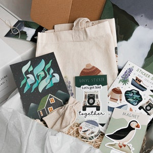 May include: A gift box filled with various items, including a tote bag, cards, stickers, and a magnet. The items feature illustrations of Icelandic landscapes, puffins, and a car. The box is labeled "Auf ins h&ouml;chste Abenteuer" and "Alles Liebe Hannah!"