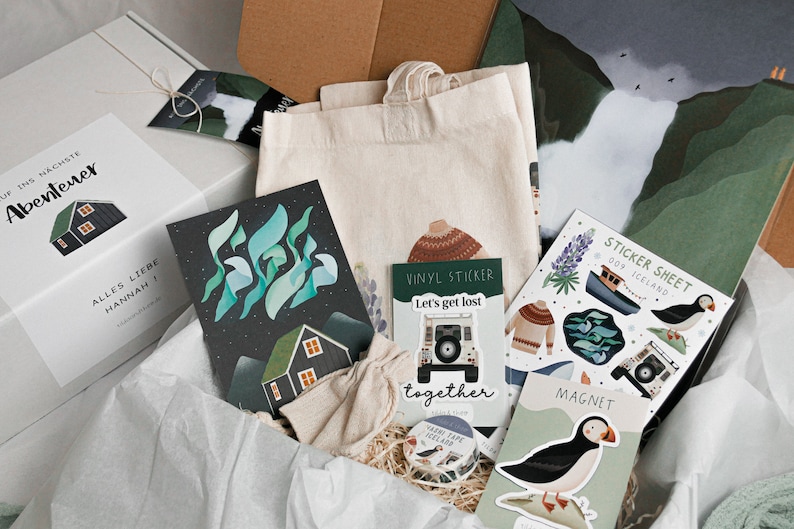May include: A gift box filled with various items, including a tote bag, cards, stickers, and a magnet. The items feature illustrations of Icelandic landscapes, puffins, and a car. The box is labeled "Auf ins h&ouml;chste Abenteuer" and "Alles Liebe Hannah!"