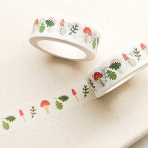 May include: Two rolls of white washi tape with a repeating pattern of red mushrooms, green leaves, and white dots. The tape is unrolled and shows the pattern.