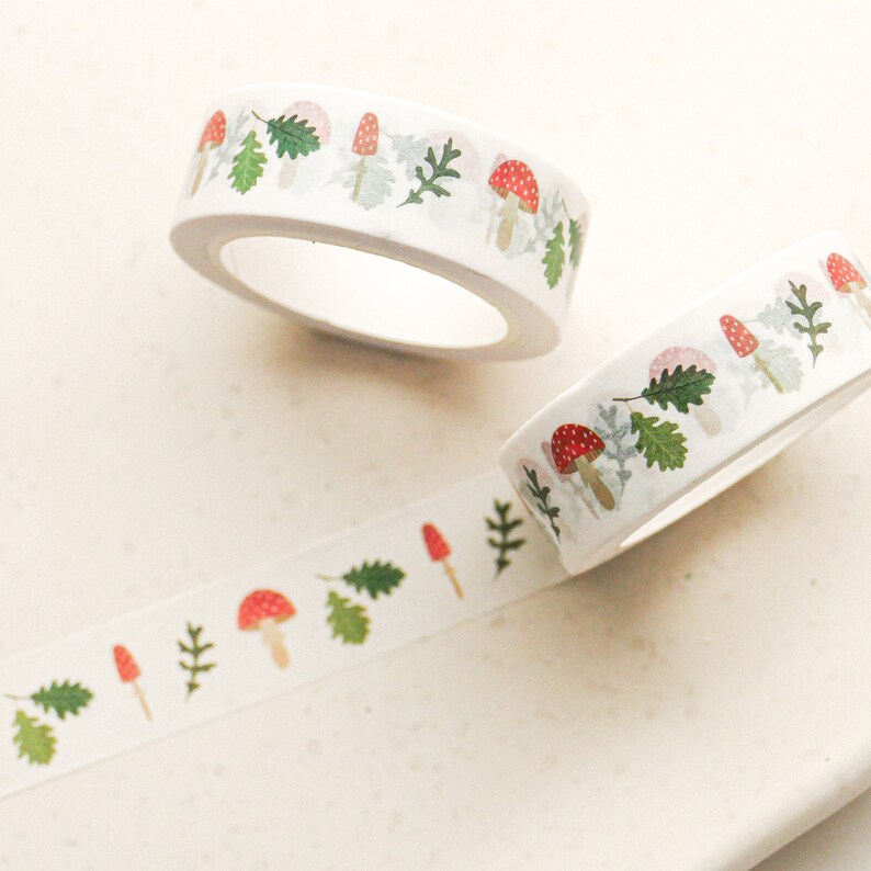 May include: Two rolls of white washi tape with a repeating pattern of red mushrooms, green leaves, and white dots. The tape is unrolled and shows the pattern.