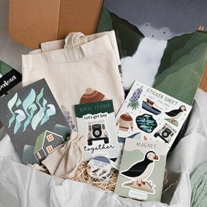 May include: A gift box filled with items themed around Iceland, including a tote bag, a sticker sheet, a magnet, a vinyl sticker, and a card with the text "Let's get lost together".