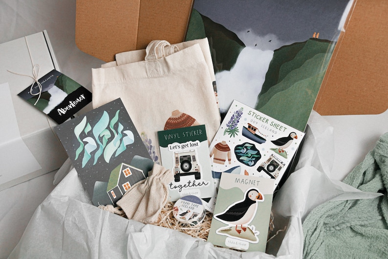 May include: A gift box filled with items themed around Iceland, including a tote bag, a sticker sheet, a magnet, a vinyl sticker, and a card with the text "Let's get lost together".