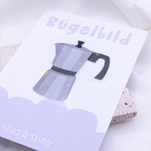 Könnte beinhalten: Eine weiße Karte mit dem Text "Bügelbild" in lila Schrift. Eine graue und schwarze Illustration einer Kaffeekanne befindet sich auf der Karte. Der Text "tilda & theo" befindet sich unten auf der Karte in lila Schrift.